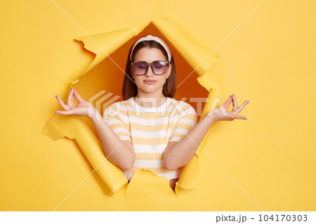 Portrait of relaxed dark haired Caucasian woman wearing striped shirt and hair band posing in yellow paper hole, relaxing, calming down, meditating, keeps eyes closed. 104170303