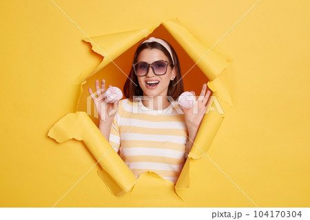Portrait of attractive young dark haired woman wearing hair band and striped t shirt standing in yellow paper hole, holding marshmallows in hands, being amazed. Portrait of attractive young dark haired woman wearing hair band and striped t shirt standing in yellow paper hole, holding marshmallows in hands, being amazed. 104170304