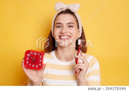 Horizontal shot of cheerful woman with dark hair, smiles toothily shows white perfect teeth, holds toothbrush and red alarm clock dressed in casual shirt poses indoors. Horizontal shot of cheerful woman with dark hair, smiles toothily shows white perfect teeth, holds toothbrush and red alarm clock dressed in casual shirt poses indoors. 104170340