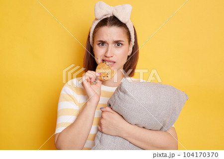 Portrait of young adult dark haired woman biting cookie eating sweets isolated on yellow background, being in bad mood, looking at camera with sad expression. Portrait of young adult dark haired woman biting cookie eating sweets isolated on yellow background, being in bad mood, looking at camera with sad expression. 104170370