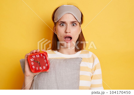 Shocked surprised woman wearing striped shirt and sleeping mask, holding pillow and red alarm clock, posing isolated over yellow background, looking at camera with open mouth and big eyes, being late. 104170450