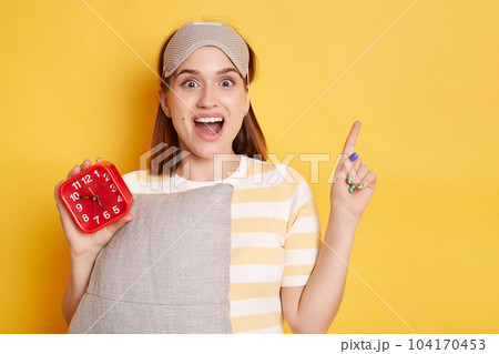 Horizontal shot of excited Caucasian woman looking at camera prepares for sleep holds soft pillow dressed in sleepmask on forehead, pointing aside with finger, showing copy space for advertisement. 104170453