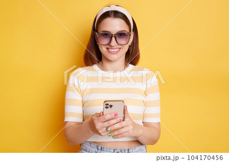 Indoor shot of attractive young woman wearing striped t-shirt standing with smart phone in hands, using cell phone, looking at camera with smile, posing isolated over yellow background. 104170456