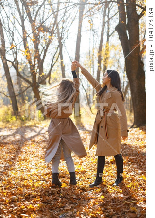 Two beautiful female friends spending time together. Two young smiling sisters walking in autumn park. Brunette and blonde girls wearing coats. 104173134