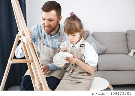Little girl with Down syndrome wearing white shirt and beige apron. Girl with her father is painting on easel together. Father helping his daughter to paint. Little girl with Down syndrome wearing white shirt and beige apron. Girl with her father is painting on easel together. Father helping his daughter to paint. 104173205