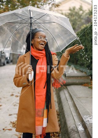 Young black woman with a long locs hairstyles standing outside with a transparent umbrella. Woman wearing brown coat, orange scarf and black hat. Woman in glasses resting outside on autumn. Young black woman with a long locs hairstyles standing outside with a transparent umbrella. Woman wearing brown coat, orange scarf and black hat. Woman in glasses resting outside on autumn. 104173283