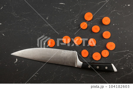 Top down view, chefs knife and small circles of sliced carrot on black marble like working board. 104182603