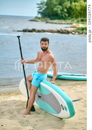 Handsome athletic man standing with paddle board on sandy beach near sea, enjoying vacation. Handsome athletic man standing with paddle board on sandy beach near sea, enjoying vacation. 104184774