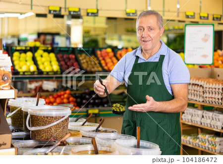 Elderly salesman showing olives in grocery store 104186472