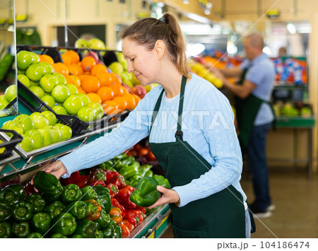 Middle-aged saleswoman putting peppers on food stall in greengrocery 104186474