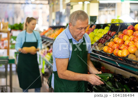 Focused mature salesman laying out small green cucumbers in supermarket 104186494