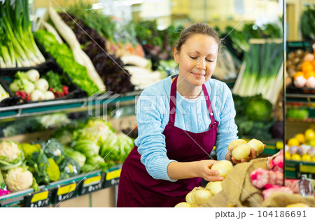 Skillful busy middle-aged female seller in uniform putting fresh onion on shelves in grocery shop 104186691