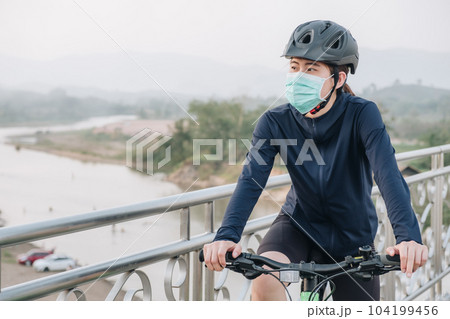 Portrait of young woman wearing a mask while riding a bicycle outdoor for protect and prevent the spread of virus. Conceptual of new normal lifestyle, everybody should wearing a mask when go outdoor. Portrait of young woman wearing a mask while riding a bicycle outdoor for protect and prevent the spread of virus. Conceptual of new normal lifestyle, everybody should wearing a mask when go outdoor. 104199456