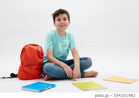Confident studio portrait of Caucasian adorable teenage boy in casual clothes, smiling looking at camera, sitting near his orange backpack and textbooks, isolated on white background. Back to school Confident studio portrait of Caucasian adorable teenage boy in casual clothes, smiling looking at camera, sitting near his orange backpack and textbooks, isolated on white background. Back to school 104201962