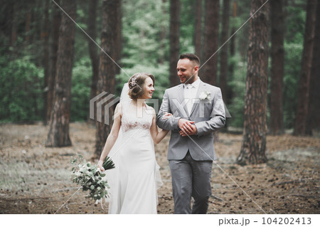 Stylish couple of happy newlyweds walking in the park on their wedding day with bouquet 104202413