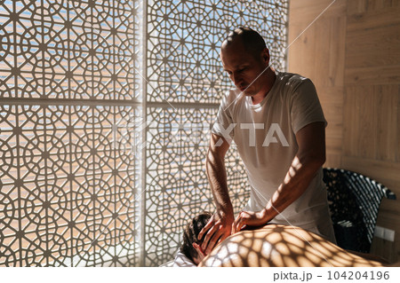 Side view of concentrated male masseur massaging neck of young sporty man lying on massage table in sunlight and beautiful shadows. Relaxation and revitalization of muscles after training. Side view of concentrated male masseur massaging neck of young sporty man lying on massage table in sunlight and beautiful shadows. Relaxation and revitalization of muscles after training. 104204196