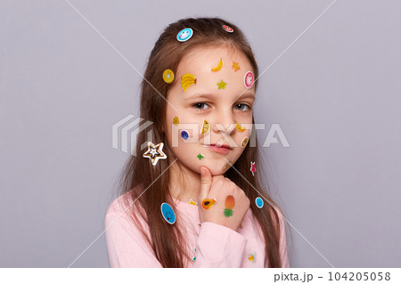 Portrait of cunning sly pensive little brown haired little girl covered with stickers posing isolated over gray background, holding chin, thinking about evil plan or prank. Portrait of cunning sly pensive little brown haired little girl covered with stickers posing isolated over gray background, holding chin, thinking about evil plan or prank. 104205058