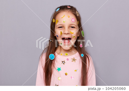 Indoor shot of excited extremely happy little brown haired little girl covered with stickers screaming with joyful expression, rejoicing posing isolated over gray background 104205060