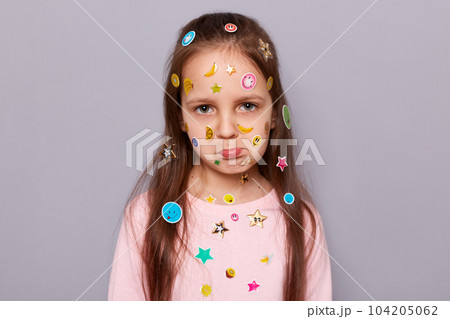 Indoor shot of sad upset little brown haired little girl covered with stickers posing isolated over gray background, feeling bored, looking at camera with pout lips. Indoor shot of sad upset little brown haired little girl covered with stickers posing isolated over gray background, feeling bored, looking at camera with pout lips. 104205062
