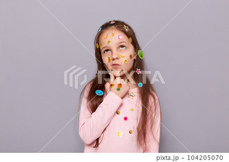 Photo of thoughtful pensive little brown haired little girl covered with stickers posing isolated over gray background, holding her chin, thinking, pondering. Photo of thoughtful pensive little brown haired little girl covered with stickers posing isolated over gray background, holding her chin, thinking, pondering. 104205070