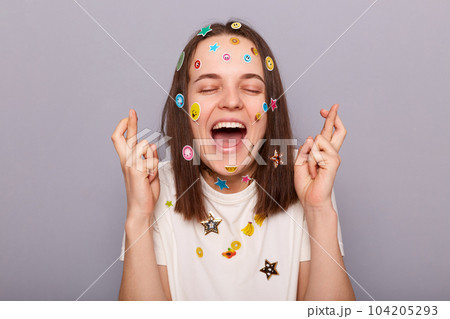 Horizontal shot of extremely happy woman covered with funny stickers posing isolated over gray background, crossing her fingers,closed eyes, praying for good luck and success. 104205293