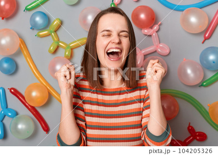 Portrait of overjoyed happy woman with brown hair wearing striped dress standing with clenched fists around multicolored inflated balloons, rejoicing, screaming. 104205326