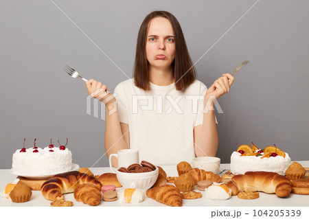 Portrait of sad stressed Caucasian woman with brown hair sitting at table isolated over gray background, looking at camera with pout lips, being upset, keeps diet, wants sweet bakery. Portrait of sad stressed Caucasian woman with brown hair sitting at table isolated over gray background, looking at camera with pout lips, being upset, keeps diet, wants sweet bakery. 104205339