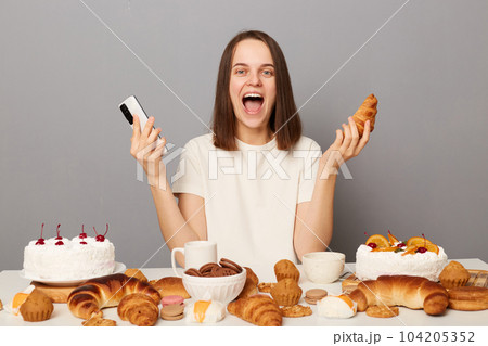 Amazed woman with brown hair sitting at table with mobile phone in hand among various desserts, has good appetite excited with variety of confectionery, tired of diets isolated over gray background 104205352