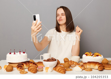 Beautiful positive woman with brown hair wearing white T-shirt sitting at table poses for selfie uses smartphone shows yummy desserts, pout lips, isolated over gray background Beautiful positive woman with brown hair wearing white T-shirt sitting at table poses for selfie uses smartphone shows yummy desserts, pout lips, isolated over gray background 104205353