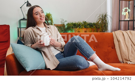 Attractive smiling young woman drinking a cup of coffee or herbal tea relaxing resting at home couch 104214792