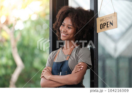 Successful african woman in apron standing coffee shop door. Happy small business owner holding tablet and working. Smiling portrait of SME entrepreneur seller business standing with copy space. Successful african woman in apron standing coffee shop door. Happy small business owner holding tablet and working. Smiling portrait of SME entrepreneur seller business standing with copy space. 104216309