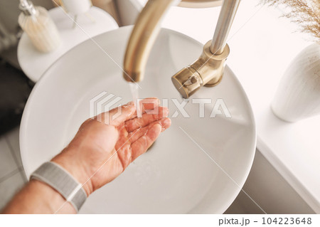 A man's hand touches the water in a beautiful sink with a golden faucet next to an oval mirror and a shelf with hand towels. Close-up of an elegant golden faucet in the bathroom sink next to stylish A man's hand touches the water in a beautiful sink with a golden faucet next to an oval mirror and a shelf with hand towels. Close-up of an elegant golden faucet in the bathroom sink next to stylish 104223648