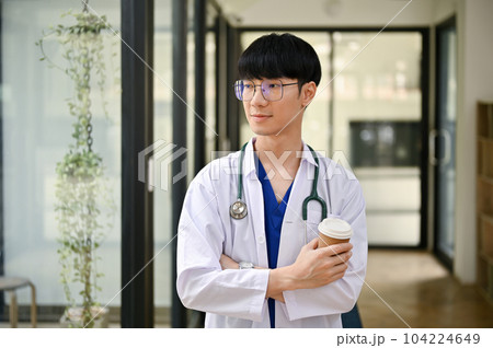 A handsome Asian male doctor in a uniform with a stethoscope stands in the hospital corridor A handsome Asian male doctor in a uniform with a stethoscope stands in the hospital corridor 104224649