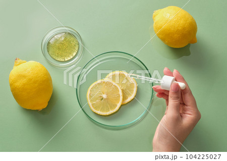 Against the pastel green background, fresh lemons decorated with petri dish containing lemon slices and essence. Female hand holding a dropper. Scene for advertising cosmetic of lemon extract 104225027