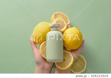 On the pastel green background, female hands holding fresh lemon and lemon slices, in the middle is green bottle unbranded mockup for design. Scene for advertising. Cosmetic of natural lemon extract 104225029