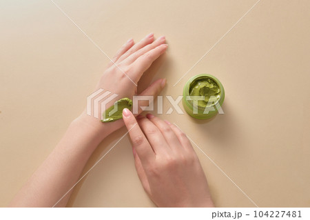 Top view of hand model applying green smear of creamy texture on her hand with a green jar beside. Light beige background Top view of hand model applying green smear of creamy texture on her hand with a green jar beside. Light beige background 104227481