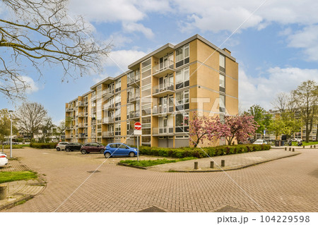 an apartment complex with cars parked on the street and trees in blooming front yard behind it, under a cloudy blue sky an apartment complex with cars parked on the street and trees in blooming front yard behind it, under a cloudy blue sky 104229598