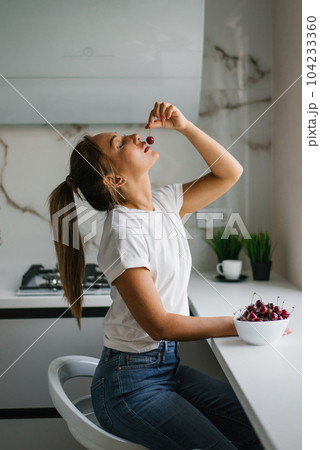 Attractive young woman eats fresh cherries, healthy berries sitting at home in the kitchen 104233360