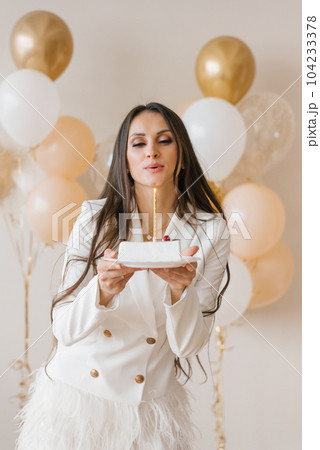 Positive woman celebrating anniversary dressed in stylish feather dress blowing out candle on birthday cake around inflated balloons at home Positive woman celebrating anniversary dressed in stylish feather dress blowing out candle on birthday cake around inflated balloons at home 104233378