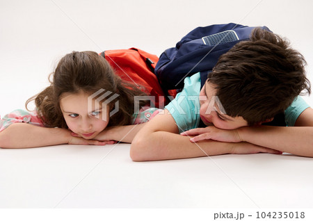 Conceptual emotional portrait of a happy little preschool girl and her brother, preteen school boy with backpacks, talking to each other in school recreation time, lying on belly over white background Conceptual emotional portrait of a happy little preschool girl and her brother, preteen school boy with backpacks, talking to each other in school recreation time, lying on belly over white background 104235018