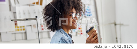 Smiling African-American lady with cup and glasses leans onto table with sewing machine in workshop Smiling African-American lady with cup and glasses leans onto table with sewing machine in workshop 104238170