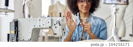 Smiling African-American seamstress shows black thread to camera sitting at workplace with sewing machine 104238171