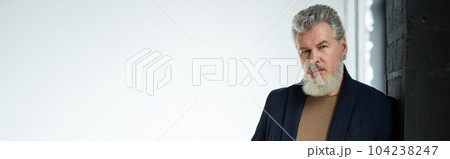 Handsome grey haired middle aged man in business casual wear looking at camera, holding smartphone, leaning on the wall while posing against white background 104238247