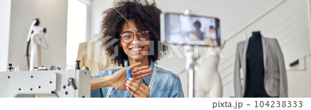African-American lady holds blue threads shooting tutorial at sewing machine in workshop 104238333