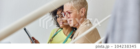 Middle aged woman with tablet sits with African-American colleague working on laptop on staircase 104238345