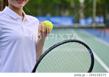 Smiling young female tennis player holding ball racket sitting on the bench at tennis court Smiling young female tennis player holding ball racket sitting on the bench at tennis court 104238575