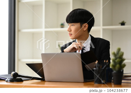 Thoughtful young businessman sitting at office desk and looking out of the window, daydreaming, visualizing future Thoughtful young businessman sitting at office desk and looking out of the window, daydreaming, visualizing future 104238677