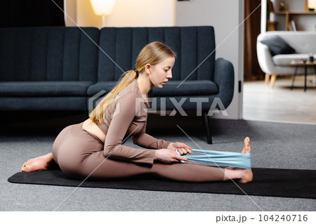 Young yogi woman practicing yoga concept, doing exercise stretching legs with elastic band, wearing sportswear bra and pants on the floor in living room 104240716