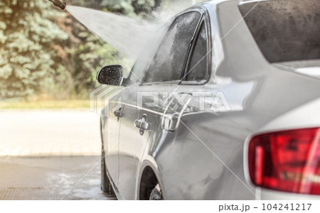 Silver car washed in self service carwash, jet water spraying from nozzle to side window, sun lit trees in background. Silver car washed in self service carwash, jet water spraying from nozzle to side window, sun lit trees in background. 104241217