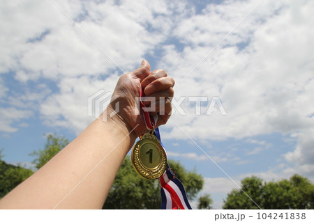 Blurred of woman hands raised and holding gold medals with Thai ribbon against blue sky background to show success in sport or business, Winners success award concept. 104241838